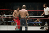 Two shirtless boxers are engaged in a match inside a boxing ring, surrounded by ropes. One boxer is wearing red shorts and the other is wearing blue shorts. A referee, dressed in a white shirt and black pants, is observing them closely. The audience is visible in the background.