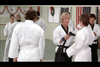 A martial arts class with several individuals wearing white gis is shown. Two people are in the foreground facing each other, with one holding a black belt. An American flag and martial arts symbol adorn the wall in the background.