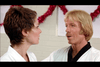 Two people, a woman with short brown hair on the left and a man with blond hair and a mustache on the right, are conversing. Both wear white martial arts uniforms. Red decorations are visible in the background.