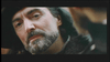 A close-up of a person with a beard and mustache, wearing a dark hat and coat, looking slightly to the side. The background is out of focus, emphasizing the person's contemplative expression. The lighting casts soft shadows on the person's face.
