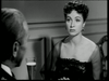 A woman with short, curly hair, wearing an elegant black dress with sheer sleeves, sits at a table. She appears to be in conversation with an older man, who is partially visible. A glass of beer is in front of the man. The image is in black and white.