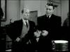 Two men wearing suits are standing indoors, engaged in conversation. The man on the left, with a bald spot, looks towards the other man, who has slicked-back hair and is holding an object in his hands. Both appear serious, in a black-and-white setting.