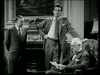 Three men are in a room. One is sitting in an armchair reading a paper, while the other two stand nearby. One man stands with his hand on his hip, and the other has his hands behind his back. They are all dressed in suits. The room has a traditional decor.