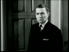A black-and-white image of a middle-aged man standing indoors, wearing a suit and tie. He has a serious expression on his face and is positioned in front of a wooden door. His hair is neatly combed, and part of a wall is visible on the right side of the image.