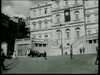 Black and white photo of a grand building with a large Nazi flag featuring a swastika hanging on its facade. The building has multiple stories, a set of grand stairs at the front, and several people and a car are visible in the courtyard.