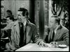 A black-and-white photo of two men in suits seated at a table, appearing to be in a restaurant. The man on the left is gesturing with his right hand, while the man on the right looks towards him. Two salt and pepper shakers are on the table.
