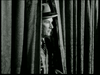 Black-and-white image of a man wearing a fedora hat and a suit, peeking through a set of heavy, dark curtains. Only his face and part of his upper body are visible as he looks intently to his right. The setting appears to be dimly lit and possibly indoors.