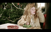 A child with long, curly hair, dressed in a beige jacket, sits at a table near a decorated Christmas tree. On the table, there are bowls filled with craft supplies, and the child is holding a piece of green foliage while looking to the side.