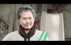 A person with gray hair and a friendly expression holds a green rolled-up poster. They are dressed warmly in a sweater and scarf. The background shows a dimly lit room with a beige curtain and some faint wall decor.