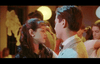 A young woman and man sharing a slow dance at a dimly lit event, gazing into each other's eyes with others dancing in the background. The ambiance is warm, with soft yellow lights illuminating the scene.