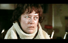 A person with short dark hair, wearing a white turtleneck sweater, appears to be in a serious or intense mood. The background is blurred and indistinct, with some objects faintly visible.
