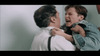 A scene from a film depicting an emotional moment where a young person, in a denim jacket, appears distressed and is grabbing an older person by the collar. The older person is wearing a white shirt and suspenders, standing against a light-colored wall.