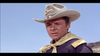 A man wearing a beige cowboy hat and a dark military-style uniform with gold trim looks into the distance against a clear blue sky. He appears to be in a desert or open plain setting.
