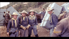 A group of soldiers wearing light blue uniforms and beige cowboy hats stand in a rugged terrain with horses and equipment boxes in the background. The scene has a historical or western setting.