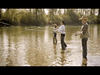 Three people are fly fishing while standing in a shallow body of water surrounded by trees. They are positioned in a line, with rods cast into the water. The scene is set in daylight with a calm, serene atmosphere.