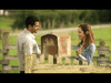 A man and a woman stand facing each other in conversation while leaning on a wooden fence. The background features a green field and trees, with a rusty mailbox visible between them. Both are casually dressed, and the atmosphere appears calm and pleasant.