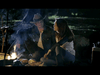 A man wearing a hat and a woman are seated close together by a campfire at night. The warm light of the fire illuminates their faces as they engage in conversation. The surrounding area is dark, with hints of trees in the background.
