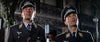 Two men in dark blue WWII-era German Luftwaffe uniforms and caps stand side by side at night, a scene from "Battle of Britain (1969)" DVD. Blurred buildings and a tree fill the background, evoking classic aerial combat drama.