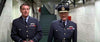 Two men in WWII-style uniforms walk down a white-walled corridor with exposed pipes and stairs, as seen in the film "Battle of Britain (1969)" DVD. Both wear dark blue jackets with medals; one has a peaked cap.