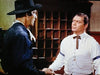 Two men in classic Western attire face off indoors, capturing the suspenseful mood of "The Bounty Hunter (1954)" DVD—one in a black hat and dark shirt, the other in a white shirt with a decorative tie, deep in serious conversation.