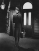 A woman in vintage clothing stands alone on a dimly lit street holding a basket, with tall buildings and a glowing streetlamp in the background—an evocative scene reminiscent of “Brief Encounter” (1945), capturing the timeless allure found in classic movies like those available on DVD.