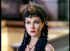 A woman in ancient-style attire adorned with an intricate blue and gold headpiece and ornate jewelry gazes intensely forward; she has fair skin, dark hair, and striking makeup evocative of the classic 1945 film “Caesar and Cleopatra” starring Claude Rains, set against a softly blurred background.