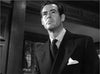 A man in a dark suit and tie with a pocket square gazes upward with a serious 1940s Hollywood look. Elegant wood paneling and decorative windows set the scene in this black-and-white image from the "Caught (1949) DVD.