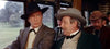 Two men in old-fashioned suits and hats sit together on a train, deep in conversation—just like a scene from the rare "Rage at Dawn (1955) DVD," with other riders and lush greenery visible through the window behind them.