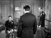 Three men in military uniforms are in an office with chairs, a wall certificate, and a large map—capturing the atmosphere of classic films like "They Were Not Divided (1950)" on DVD. One sits while two stand, one facing away and the other behind a desk.