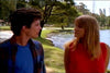 A young man and woman stroll by a lakeside on a sunny day, discussing their favorite classic movies like "Crossing the Line (1990)" on DVD, surrounded by grass, trees, and engaging conversation.