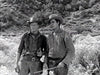 Two men in cowboy hats stand outdoors in a rugged, black-and-white scene inspired by classic films. Hills and bushes frame the background as they hold tools—evoking the spirit of The Yellow Tomahawk (1954) DVD.