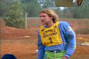 A man with long blond hair and a mustache, wearing a blue shirt and yellow GAMEX CHALLENGE 1 vest, stands on a dirt motocross track outdoors in "Crossing the Line (1990)" DVD. Trees and fencing appear in the background.