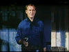 A man in a plaid shirt stands outside by a shadowy wooden structure, holding a handgun and looking serious—reminiscent of scenes from the "Rage at Dawn (1955) DVD.