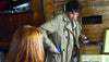 A man in a beige trench coat, holding a phone and blue object by a wooden wall, has a Dear Prudence (2009) DVD in his pocket. Nearby, a woman with reddish-brown hair sits with shelves and books behind them.