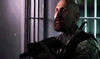 A bald bearded man in military gear stands tensely by metal jail bars in a dim setting—an intense moment from the rare, hard-to-find movie collection featuring Jurassic City (2015) DVD.