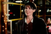 A woman with dark hair and a brown blazer smiles indoors, surrounded by soft string lights and blurred figures—evoking the cozy, classic movie atmosphere found in "When Friendship Kills (1996)" on DVD.