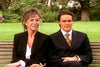 A woman in glasses and a man in a suit sit together on a park bench, both smiling gently—evoking scenes from rare films like "A Most Deadly Family (2001) DVD," with lush trees and greenery behind them.