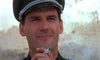 A man in uniform and cap smiles slightly with a cigarette against a blurred outdoor backdrop—evoking rare war movie scenes as seen in Wheels of Terror (1987) DVD.