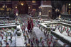 A man and woman in red walk through a snowy courtyard filled with colorful elves and twinkling lights, with a decorated building behind—evoking scenes from "The Christmas Chronicles 2 (2020) DVD.