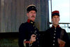 Two men in dark military uniforms and hats stand outside, one with a mustache holding a pistol, the other serious—scene from "March or Die (1977)" DVD—set against a wooden wall backdrop.