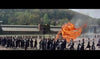 A large group in dark uniforms marches by a burning wooden structure on a field, with smoke rising and trees and a long building behind—evoking classic scenes from The Long Gray Line (1955) DVD.