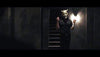 A person in a horned mask and dark clothes stands in a dim hallway by the stairs, lit by one wall lamp—evoking chilling moments from The Black Phone (2021) DVD.