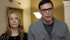 A woman with long blonde hair and a necklace stands beside a man in glasses and a gray sweater, both serious—indoors with art behind them, they resemble characters from "A Murder of Innocence (2018)" DVD.