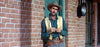 A man in a cowboy hat and vest leans against a brick wall outside, hands clasped and gun belt at his waist, looking at the camera—resembling classic films like The Oklahoman (1957) DVD.