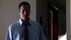 A man in a light blue shirt and patterned tie stands in dim light, looking concerned—like a scene from the "Black Widower (2006) DVD," with shadows from a nearby doorway partially illuminating him.