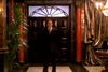 A man in a dark suit stands before grand double doors with ornate glass, red marble columns, and lush plants—reminiscent of the rare, hard-to-find film A Most Deadly Family (2001) DVD.