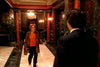 A woman in an orange top, patterned jacket, and glasses—evoking Mary Tyler Moore—walks her dog toward a suited man in an ornate hallway, as seen on the "A Most Deadly Family (2001) DVD.