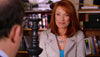 A red-haired woman in a light jacket and blue top smiles while seated in an office or library, with shelves of books and DVDs—including a Dear Prudence (2009) DVD—behind her. A blurred figure appears in the foreground.