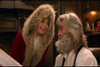 A woman in a red coat with a fur-lined hood smiles at a man with gray hair, a beard, and glasses as they sit together in a cozy wood-paneled room filled with rare movies, featuring The Christmas Chronicles (2018) DVD.