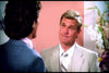 A man in a light beige suit and striped tie smiles while talking to another man in a blue suit, both in a flower-filled room—reminiscent of scenes from The Cartier Affair (1984) DVD.